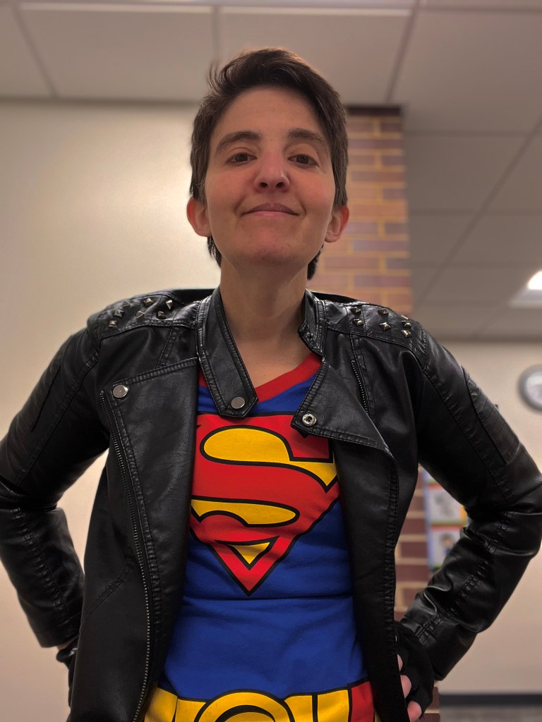 Current image: A picture of the Author (AuthorLibrarianRachel) Rachel Stine dressed as Kon-El Superboy, wearing a black pleather jacket, superman t-shirt, and with their hands on their hips
