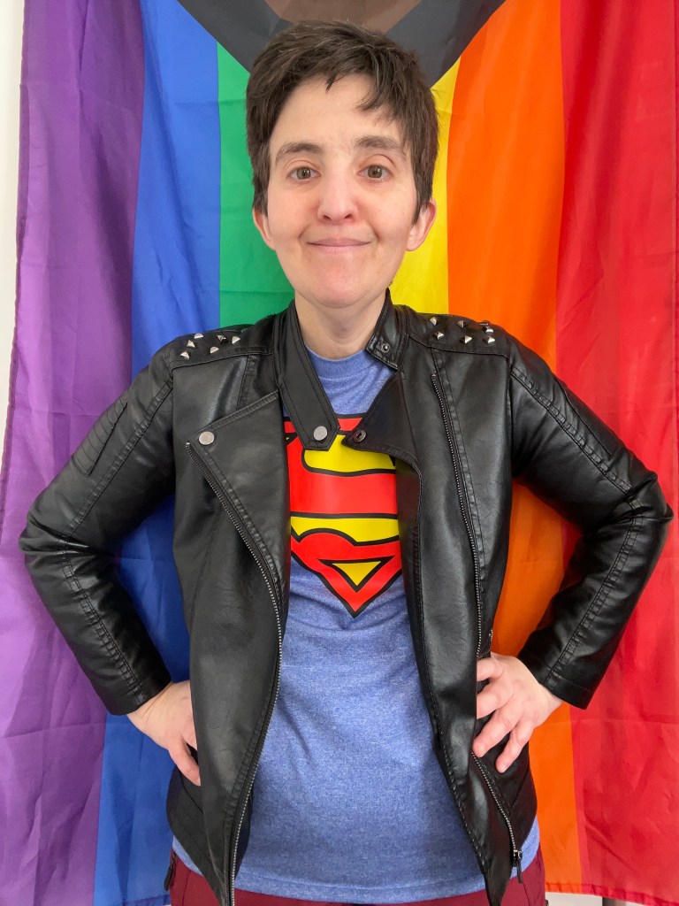Photo of the author (AuthorLibrarianRachel) Rachel A. Stine. They are dressed as Kon-El Superboy and standing in front of the progress pride flag.